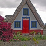 The colorful traditional stone houses with triangular-shaped thatched roofs. They have white-painted walls, red doors, and windows with blue trim.