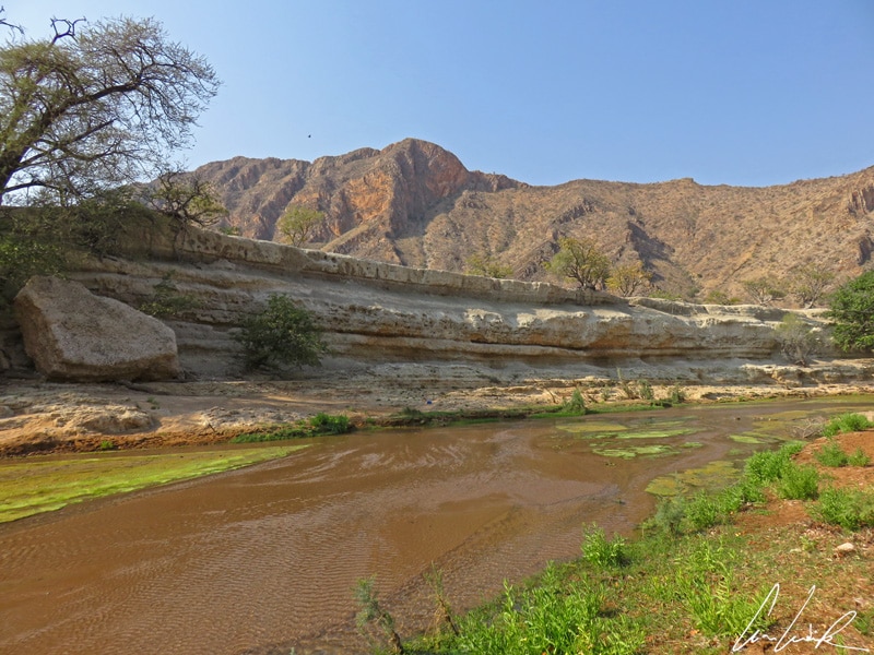 In the canyon, a shallow river flows. Vegetation is abundant on both sides of the shoreline.