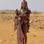 A Himba woman carries her young child in her arms. Its head is covered with a headdress, the Erembe symbolizing the ears of a cow.
