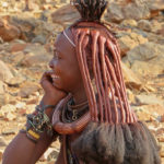 A smiling Himba woman proudly wears the Erembe, this goatskin headdress with long braids coated with clay