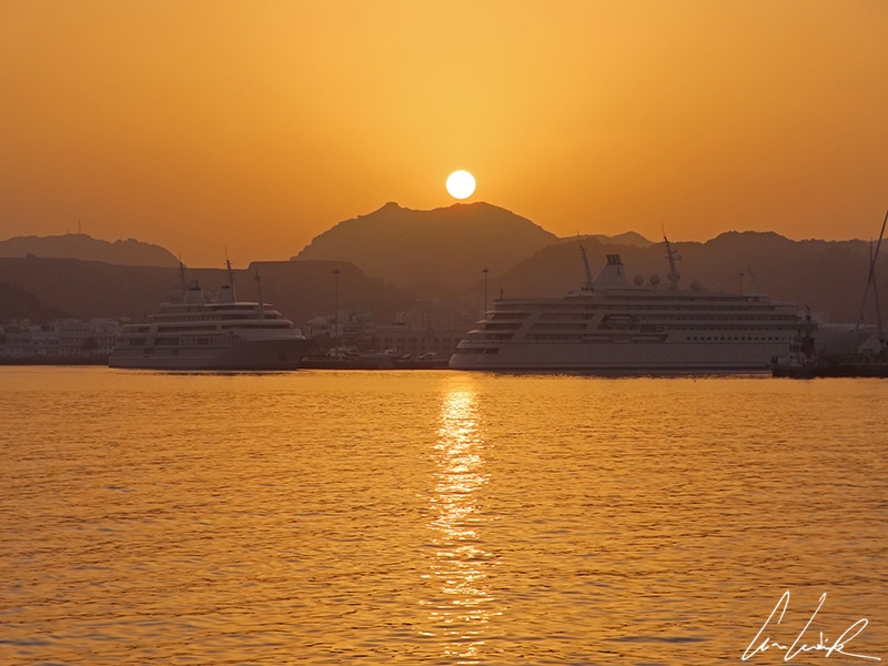 As the sun goes down behind the mountains, you can admire the two floating residences of the Sultan glisten in the Muscat harbor.