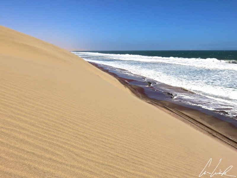 Gigantic sand dunes lie along the ocean as far as the eye can see. The desert is dressed in shimmering golden colours contrasting with the blue-green of the ocean.