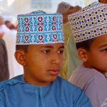 These two young children are wearing the traditional round embroidered hat: the kumma. It matches their dishdasha: blue for one and purple for the other.