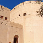 Nizwa Fort is a mixture of a fortified tower and a castle. The fort has an imposing structure with high ochre-colored walls that contrast with the blue sky.