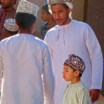 At the Nizwa cattle market men in dishdasha wearing the traditional kumma or mussar are accompanied by their young sons.