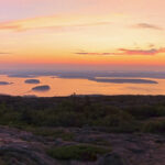 From the top of Cadillac Mountain, the view of Frenchman Bay is magnificent at dawn. The sky is colored orange.