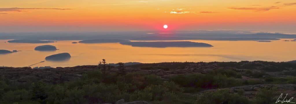 The sunrise over Acadia National Park colors the sky with orange tones. The ocean and the Porcupine Islands become visible.