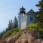 The Bass Harbor Lighthouse is a 26-foot white cylindrical tower. The black lantern at its top emits a red light.