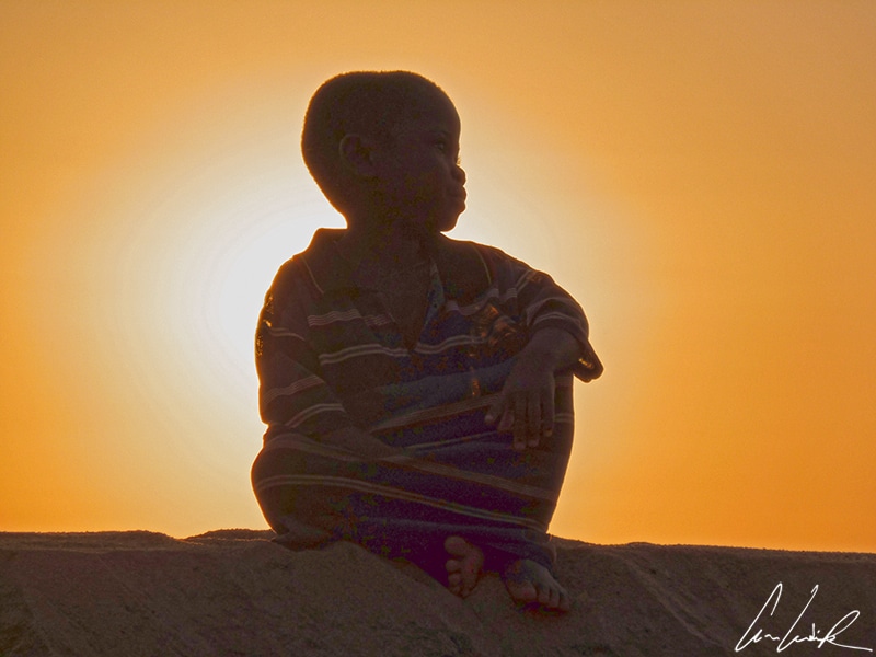 A child sitting on the banks of the Tsiribihina River at sunset in Madagascar. The Tsiribihina River is wide and quiet. The river takes its time and traces its meanders in a beautiful landscape. Nothing but silence and nature around you.