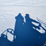 Sur la motoneige, un passager et le pilote dont les ombres se reflètent sur la neige.
